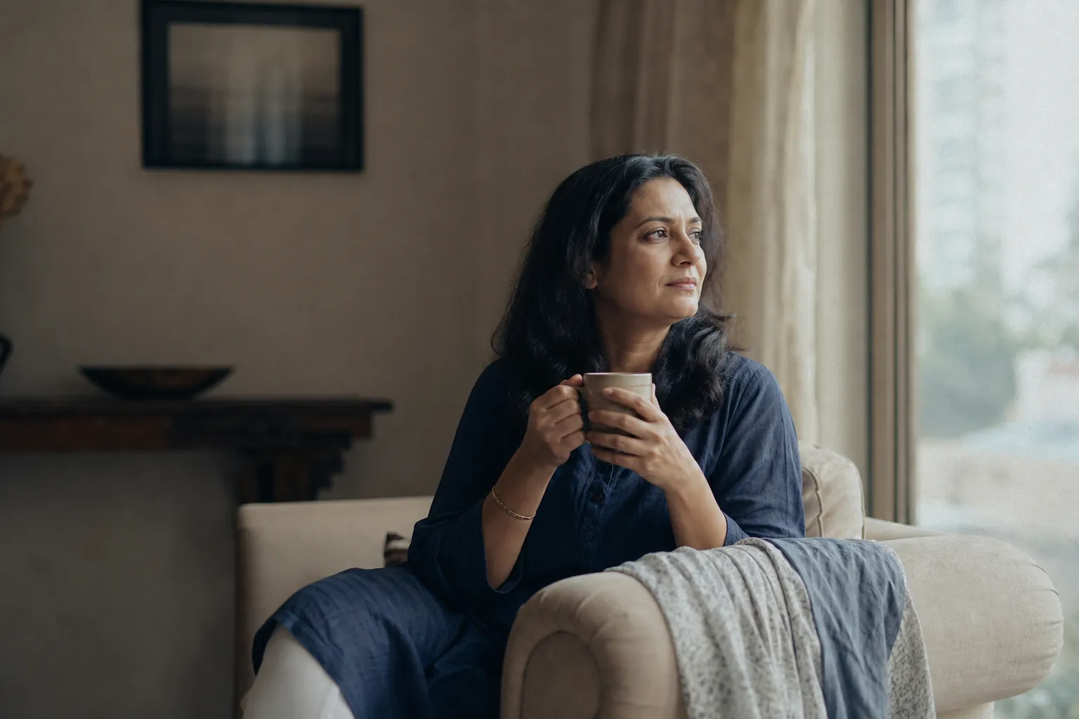 A woman sitting calmly by a window in soft morning light, holding a mug and looking thoughtfully into the distance