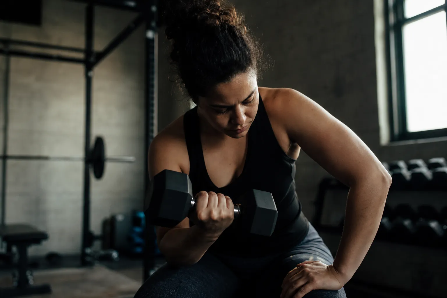 A person mid-set lifting a dumbbell in a raw concrete gym with hard directional light