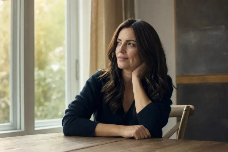 A woman sitting at a sunlit table, resting her chin on her hand and looking thoughtful as she considers a health decision